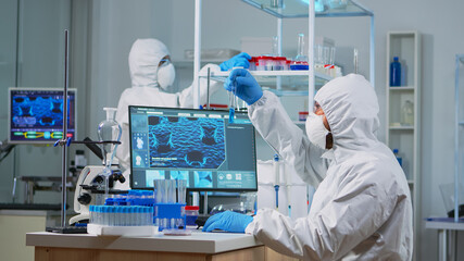 Wall Mural - Neurologist with ppe suit working at vaccine development in equipped laboratory typing on pc. Team examining virus evolution using high tech for research in treatment development against covid19