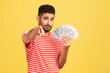 © khosrork - Self confident rich man with beard in striped t-shirt holding fan of dollars and pointing finger at camera with serious expression, easy profit. Indoor studio shot isolated on yellow background