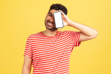 Happy cheerful bearded man in red striped t-shirt holding and showing smartphone with white empty display, looking at camera with toothy smile. Indoor studio shot isolated on yellow background