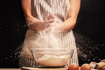 Female baker slapping hands and splashing flour while making dough for a loaf of bread with an apron. Natural homemade ingredients. Eggs and roller visible in the background.