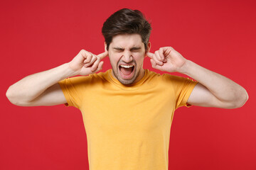 Young irritated grumpy angry nervous unshaved caucasian man 20s wearing casual yellow t-shirt closed eyes cover ears with fingers shouting screaming isolated on red color background studio portrait.