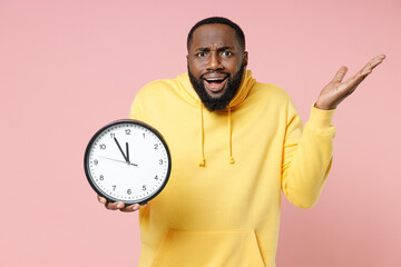 Wall Mural - Perplexed young african american man 20s wearing casual yellow streetwear hoodie standing holding clock spreading hands looking camera isolated on pastel pink color wall background studio portrait.