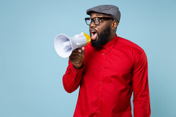 Wall Mural - Shocked amazed young bearded african american man 20s wearing casual red shirt eyeglasses cap standing screaming in megaphone looking aside isolated on pastel blue color background studio portrait.