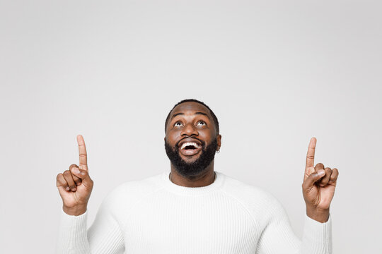Excited funny cheerful young african american man 20s wearing casual basic sweater standing pointing index fingers up on mock up copy space isolated on white color wall background studio portrait.