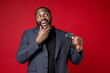 © ViDi Studio - Pensive young african american business man 20s wearing classic jacket suit standing put hand prop up on chin looking up hold credit bank card isolated on bright red color background studio portrait.