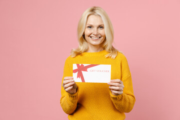 Smiling elderly gray-haired blonde woman lady 40s 50s years old wearing yellow casual sweater standing hold gift certificate looking camera isolated on pastel pink color background studio portrait.