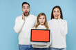 © ViDi Studio - Shocked young parents mom dad with child kid daughter teen girl in sweaters hold laptop computer with blank empty screen put hands on cheeks isolated on blue background. Family day parenthood concept.