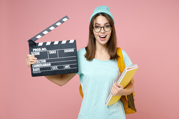 Cheerful young woman student in blue t-shirt hat glasses backpack hold notebook classic black film making clapperboard isolated on pink background. Education in high school university college concept.