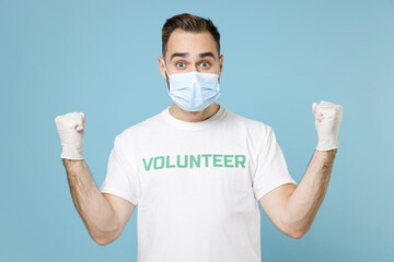 Wall Mural - Excited man in volunteer t-shirt gloves face mask to safe from coronavirus virus covid-19 doing winner gesture isolated on blue background. Voluntary free work assistance aid help support concept.