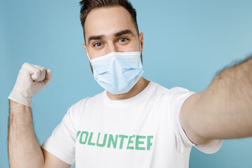 Wall Mural - Close up of man in volunteer t-shirt mask to safe from coronavirus covid-19 doing selfie on mobile phone winner gesture isolated on blue background. Voluntary work assistance aid help support concept.