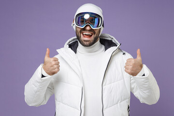 Wall Mural - Excited skier man in white windbreaker jacket ski goggles mask showing thumb up going to spend extreme weekend winter season in mountains isolated on purple background. People lifestyle hobby concept.