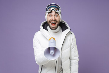 Wall Mural - Excited skier man in white windbreaker jacket ski goggles mask screaming in megaphone spend extreme weekend winter season in mountains isolated on purple background. People lifestyle hobby concept.