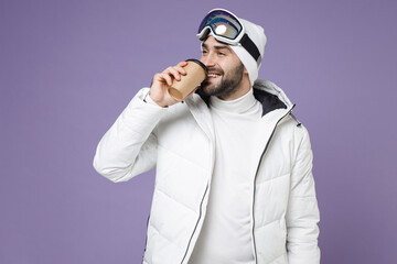 Wall Mural - Funny skier man in white windbreaker jacket ski goggles mask hold paper cup coffee tea drinking spend extreme weekend winter in mountains isolated on purple background. People lifestyle hobby concept.