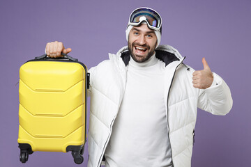 Wall Mural - Cheerful traveler tourist skier man in white jacket ski goggles mask hold suitcase showing thumb up spend weekend winter in mountains isolated on purple background. People lifestyle hobby concept.