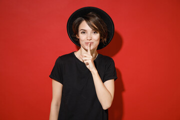 Secret young brunette woman 20s years old wearing casual basic black t-shirt hat standing saying hush be quiet with finger on lips shhh gesture isolated on bright red color background studio portrait.