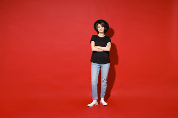 Full length of smiling young brunette woman 20s years old wearing casual basic black t-shirt hat standing holding hands crossed looking camera isolated on bright red color background studio portrait.