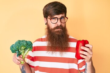 Sticker - Handsome young red head man with long beard holding broccoli and red pepper skeptic and nervous, frowning upset because of problem. negative person.