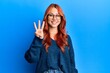 Young beautiful redhead woman wearing casual sweater and glasses over blue background showing and pointing up with fingers number three while smiling confident and happy.