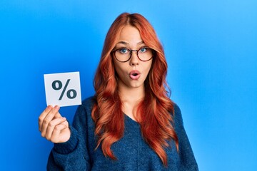 Young beautiful redhead woman holding percentage symbol scared and amazed with open mouth for surprise, disbelief face