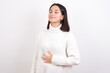 Young brunette woman wearing white knitted sweater against white background touches tummy, smiles gently, eating and satisfaction concept.
