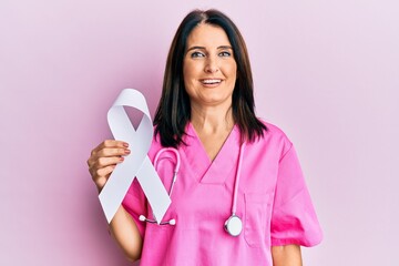 Canvas Print - Middle age brunette doctor woman holding white ribbon looking positive and happy standing and smiling with a confident smile showing teeth