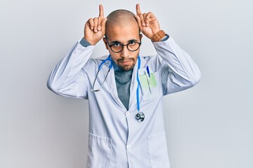 Poster - Hispanic adult man wearing doctor uniform and stethoscope doing funny gesture with finger over head as bull horns