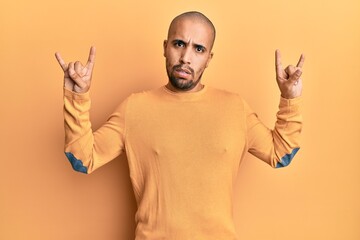 Sticker - Hispanic adult man doing rock gesture over yellow background in shock face, looking skeptical and sarcastic, surprised with open mouth