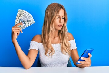 Sticker - Beautiful blonde young woman holding dollars and using smarpthone relaxed with serious expression on face. simple and natural looking at the camera.