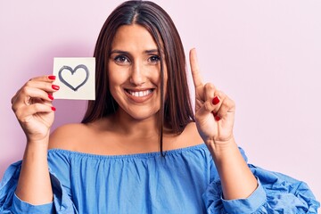 Sticker - Young beautiful brunette woman holding reminder with heart smiling with an idea or question pointing finger with happy face, number one