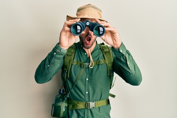 Sticker - Handsome man with beard wearing explorer hat looking through binoculars in shock face, looking skeptical and sarcastic, surprised with open mouth