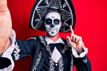 Sticker - Young man wearing mexican day of the dead costume make selfie by the camera surprised with an idea or question pointing finger with happy face, number one