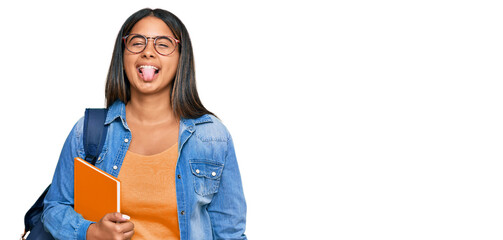 Poster - Young latin girl wearing student backpack and holding books sticking tongue out happy with funny expression. emotion concept.