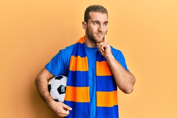 Wall Mural - Young caucasian man football hooligan cheering game holding ball serious face thinking about question with hand on chin, thoughtful about confusing idea