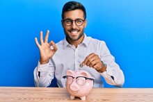 Handsome Hispanic Man Holding Piggy Bank With Glasses Doing Ok Sign With Fingers, Smiling Friendly Gesturing Excellent Symbol