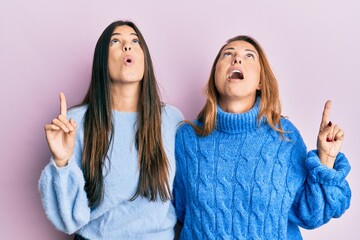 Poster - Hispanic family of mother and daughter wearing wool winter sweater amazed and surprised looking up and pointing with fingers and raised arms.