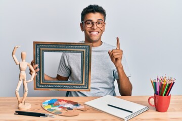 Sticker - Young handsome african american man painter sitting at art studio holding empty frame smiling with an idea or question pointing finger with happy face, number one