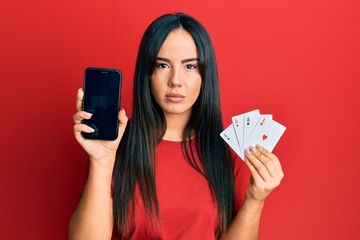 Wall Mural - Young beautiful hispanic girl holding smartphone showing screen and poker cards relaxed with serious expression on face. simple and natural looking at the camera.