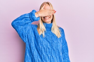 Poster - Young blonde girl wearing wool winter sweater smiling and laughing with hand on face covering eyes for surprise. blind concept.