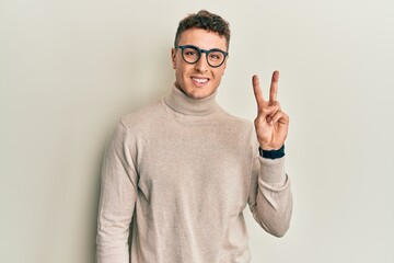 Hispanic young man wearing casual turtleneck sweater smiling looking to the camera showing fingers doing victory sign. number two.