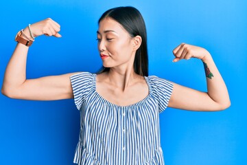 Poster - Young chinese woman wearing casual striped t-shirt showing arms muscles smiling proud. fitness concept.