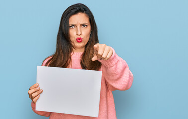 Beautiful hispanic woman holding blank empty banner pointing with finger to the camera and to you, confident gesture looking serious