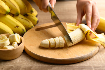 Hand holding kitchen knife and cutting ripe banana fruit on wooden cutting board for cooking