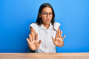 Sticker - Beautiful hispanic woman wearing casual clothes sitting on the table moving away hands palms showing refusal and denial with afraid and disgusting expression. stop and forbidden.