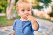 Sad little boy putting fingers on mouth touching gums because toothache at the park on a sunny day. Beautiful blonde hair male toddler in pain for new baby teeth outdoors