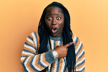 Sticker - Young black woman with braids wearing casual winter sweater surprised pointing with finger to the side, open mouth amazed expression.