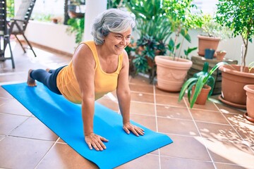 Middle age woman with grey hair smiling happy doing exercise and stretching on the terrace at home