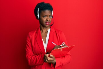 Sticker - Young african american girl wearing call center agent headset writing on clipboard clueless and confused expression. doubt concept.