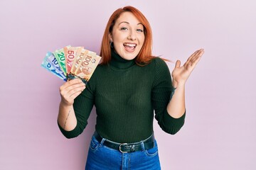 Wall Mural - Beautiful redhead woman holding canadian dollars celebrating achievement with happy smile and winner expression with raised hand