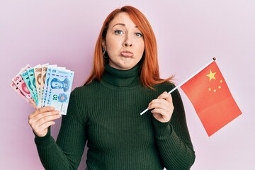 Sticker - Beautiful redhead woman holding yuan chinese banknotes and china flag depressed and worry for distress, crying angry and afraid. sad expression.