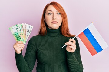 Poster - Beautiful redhead woman holding russian ruble banknotes and russia flag relaxed with serious expression on face. simple and natural looking at the camera.